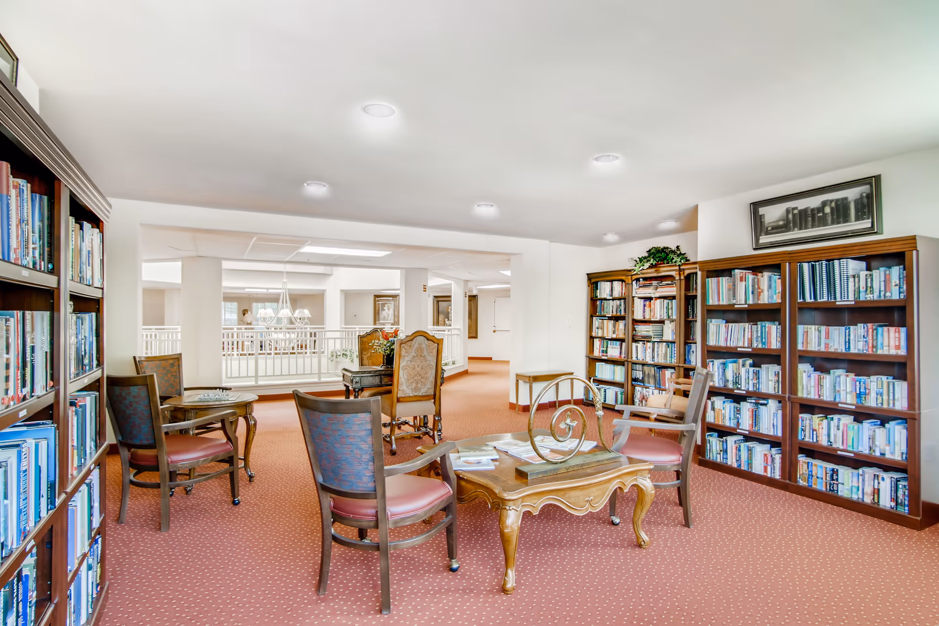Bright communal library/reading room with bookshelves, tables and chairs on red carpeting.