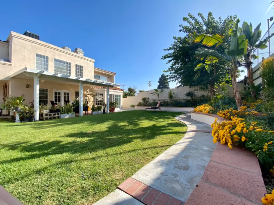 A sunny outdoor garden area with a well-maintained green lawn, a curved concrete pathway, and vibrant yellow flowers along the right side. The background features a two-story beige building with large windows and a covered patio area with potted plants and outdoor furniture. Trees and shrubs surround the garden, and the sky is clear and blue.