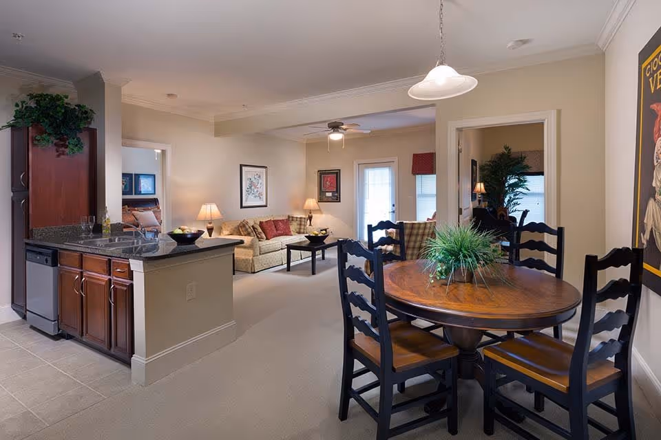 Interior view of a senior living facility showing a combined kitchen, dining, and living area. The kitchen has dark wood cabinets, a granite countertop, and a dishwasher. The dining area features a round wooden table with four chairs and a green plant centerpiece. The living room has a beige sofa with red and patterned cushions, a coffee table, framed artwork on the walls, and lamps providing warm lighting. There is a ceiling fan and a door leading outside in the background.