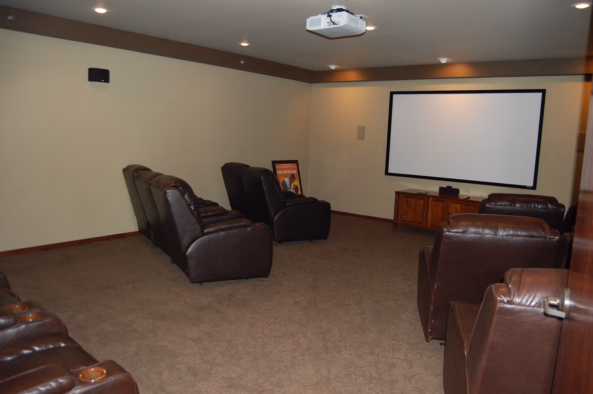 A small theater room with several brown leather recliner chairs arranged in rows facing a large white projection screen mounted on a beige wall. There is a projector mounted on the ceiling and a wooden cabinet below the screen. The room has carpeted flooring and recessed lighting in the ceiling.