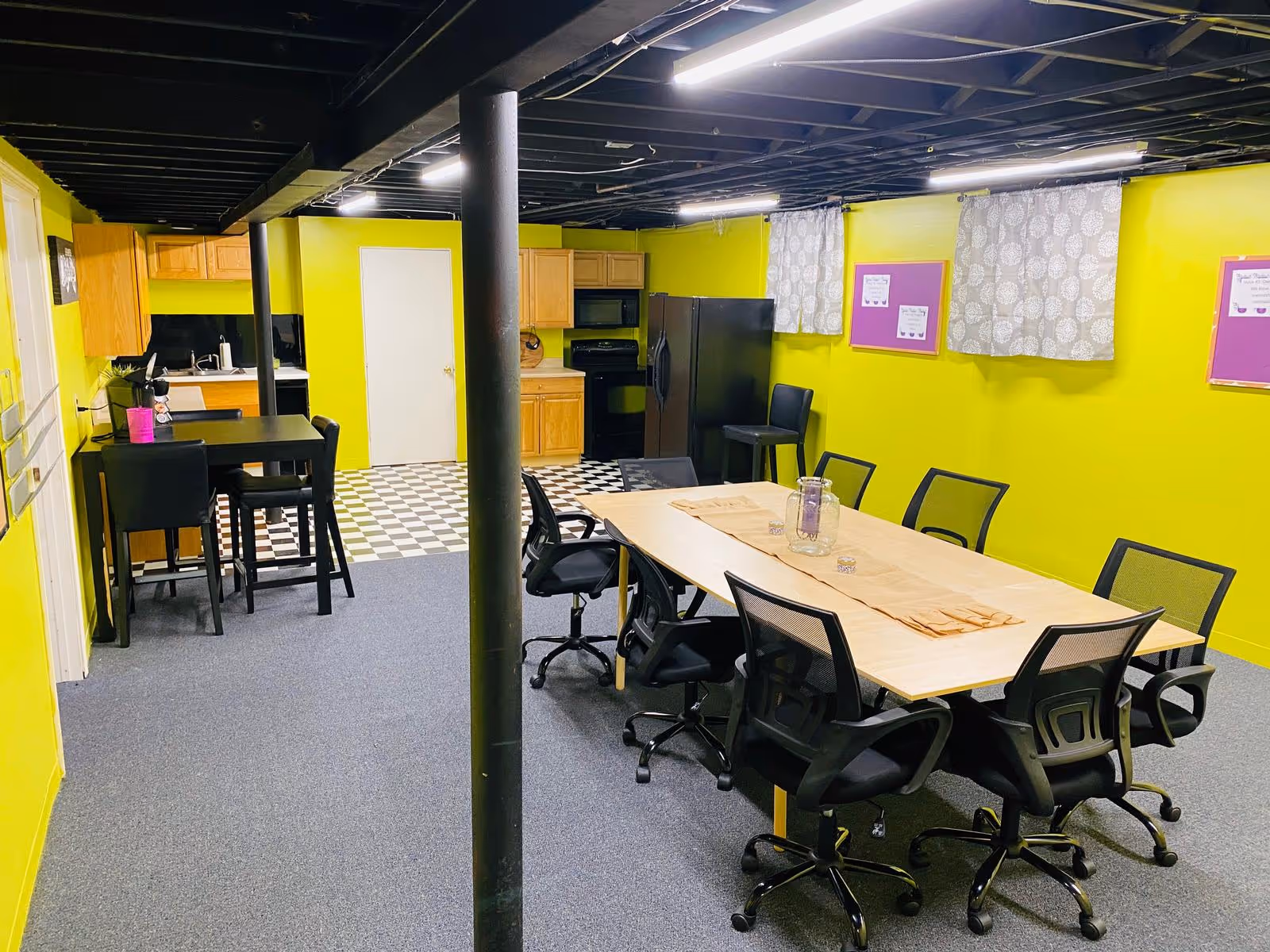 Interior room with bright yellow-green walls featuring a kitchen area with wooden cabinets, a black refrigerator, stove, and microwave. There is a checkered black and white floor in the kitchen section and carpeted flooring in the rest of the room. A large rectangular table with eight black office chairs is in the foreground, and a smaller table with two high chairs is near the kitchen. The ceiling is black with exposed beams and fluorescent lighting. Two windows with patterned curtains and bulletin boards are on the far wall.