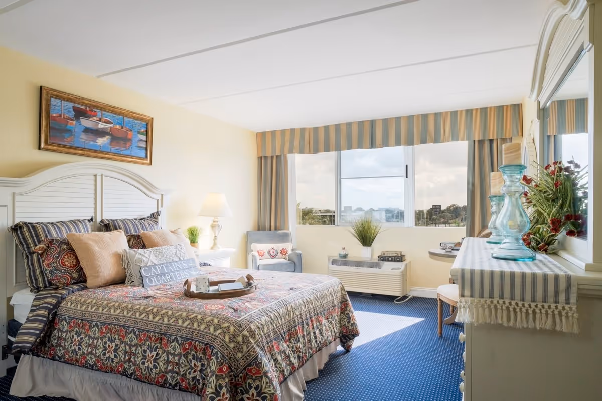 A bright and cozy bedroom with a large bed covered in a patterned quilt and multiple pillows, including one with the text 'CAPTAINS QUARTERS'. The room features a window with striped curtains, a blue armchair with a decorative pillow, a white nightstand with a lamp and plant, and a dresser with a striped runner, glass candle holders, and a floral arrangement. A framed painting of boats hangs above the bed.