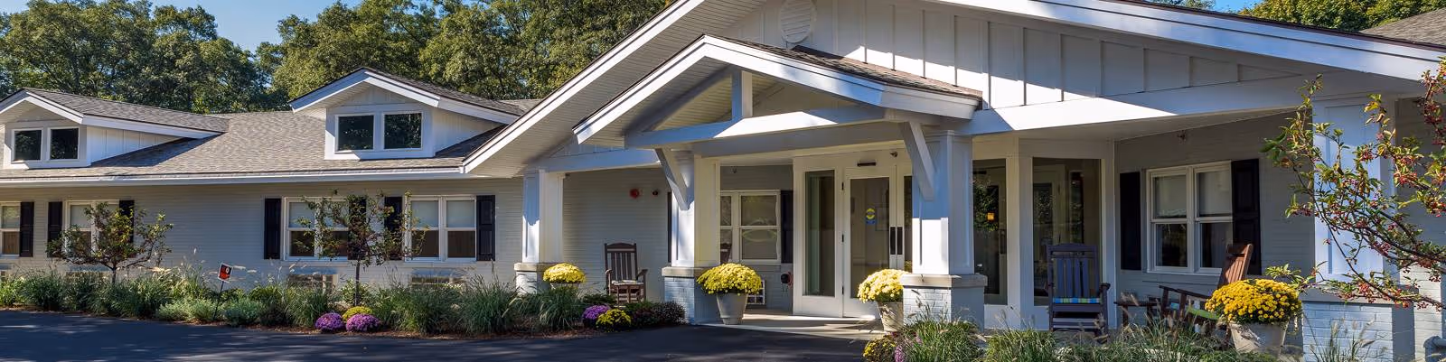 Exterior view of a single-story building with white siding and a covered entrance supported by white beams. The building has multiple windows with black shutters and dormer windows on the roof. There are potted yellow flowers and shrubs along the front, with a paved driveway in front of the entrance.