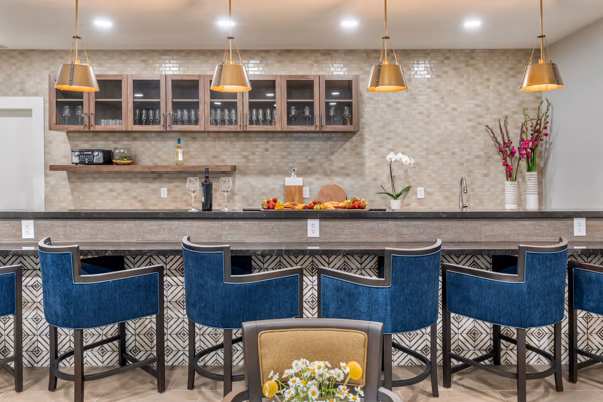 A modern kitchen area with a long counter featuring five blue upholstered bar stools. Above the counter hang four pendant lights with gold shades. Behind the counter, there are wooden cabinets with glass doors displaying glassware, a floating shelf with a radio and a bottle of wine, and a countertop with wine glasses, a bottle of red wine, a cheese and fruit platter, a cutting board, and decorative flowers in vases. The backsplash is made of beige tiles, and the floor has a patterned tile design.