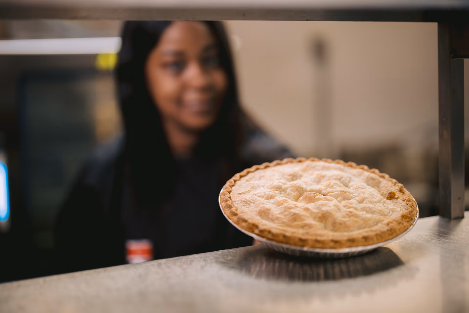 A freshly baked pie resting on a metal counter with a blurred person standing in the background.