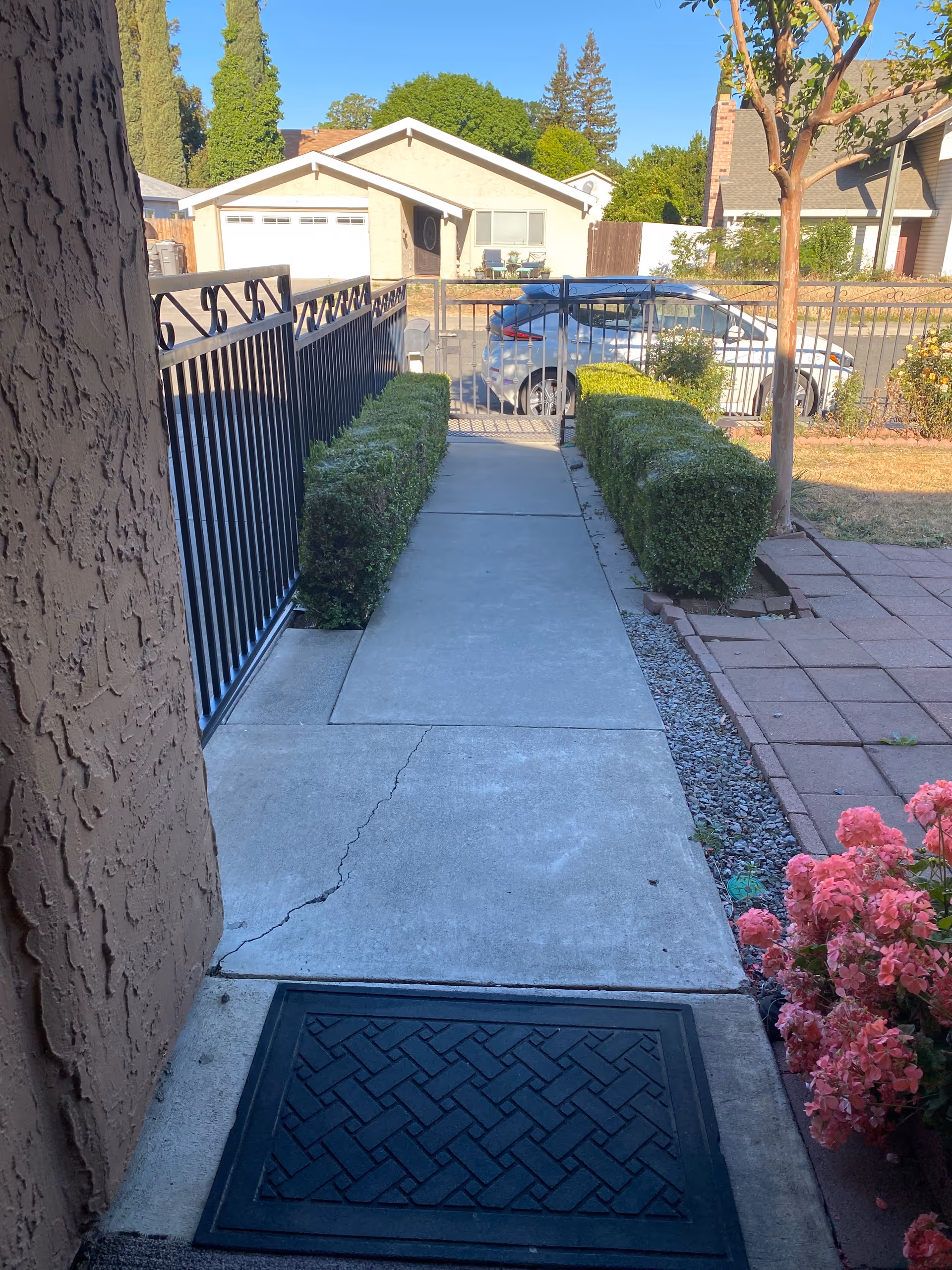 View of a concrete walkway leading from a house entrance with a black doormat, flanked by trimmed green bushes on both sides. A black metal fence and gate are visible at the end of the walkway, with a silver car parked on the street beyond. There are houses and trees in the background under a clear sky.