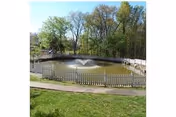 A small pond with a water fountain in the center, surrounded by a white picket fence. Trees with green foliage are visible in the background under a clear sky.