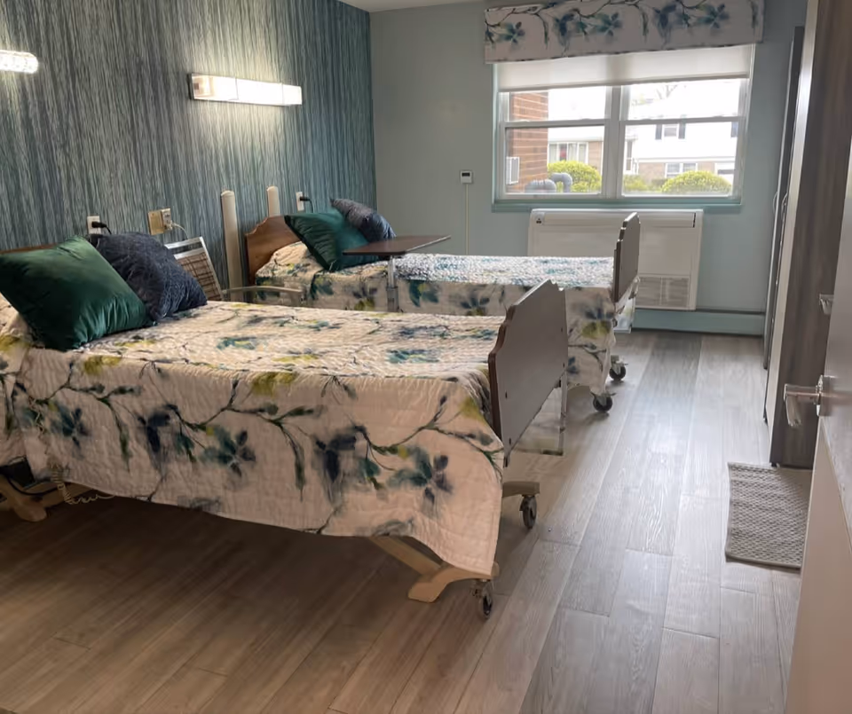 Two hospital-style beds with floral bedding and green pillows in a bright, accessible bedroom with a window and laminate floors.
