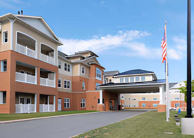 Exterior view of a multi-story senior living facility building with balconies, an entrance canopy, a driveway, and an American flag on a flagpole under a partly cloudy blue sky.