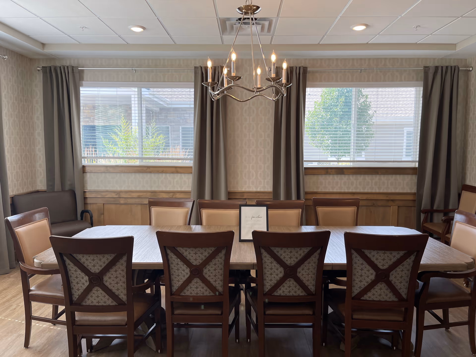 A dining room with a long rectangular wooden table surrounded by ten chairs with cushioned seats and backs. The room has two large windows with closed blinds and gray curtains, beige patterned wallpaper, wood paneling on the lower walls, and a modern chandelier hanging above the table. There is a small framed sign placed on the table.