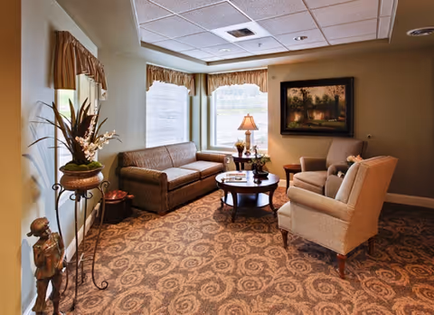 Sunlit common sitting area with a sofa, two armchairs around a coffee table, lamp, and decorative plants and artwork.