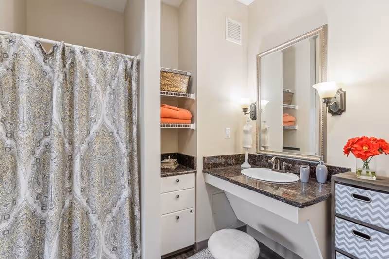 A bathroom with a patterned shower curtain on the left, a countertop with a sink and faucet, a large framed mirror above the sink, and two wall-mounted light fixtures. There are shelves with orange towels and a basket, a small white stool under the counter, and a cabinet with a chevron pattern drawer holding a vase with red flowers.