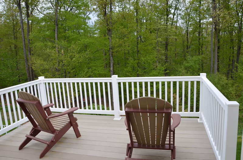 Two wooden Adirondack chairs on a deck with white railing overlooking a green forest with tall trees.
