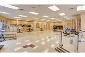 A spacious rehabilitation or physical therapy room with various exercise and therapy equipment including parallel bars, therapy tables, and chairs. The room has a tiled floor with a pattern, bright overhead lighting, and beige walls with multiple doors and windows.