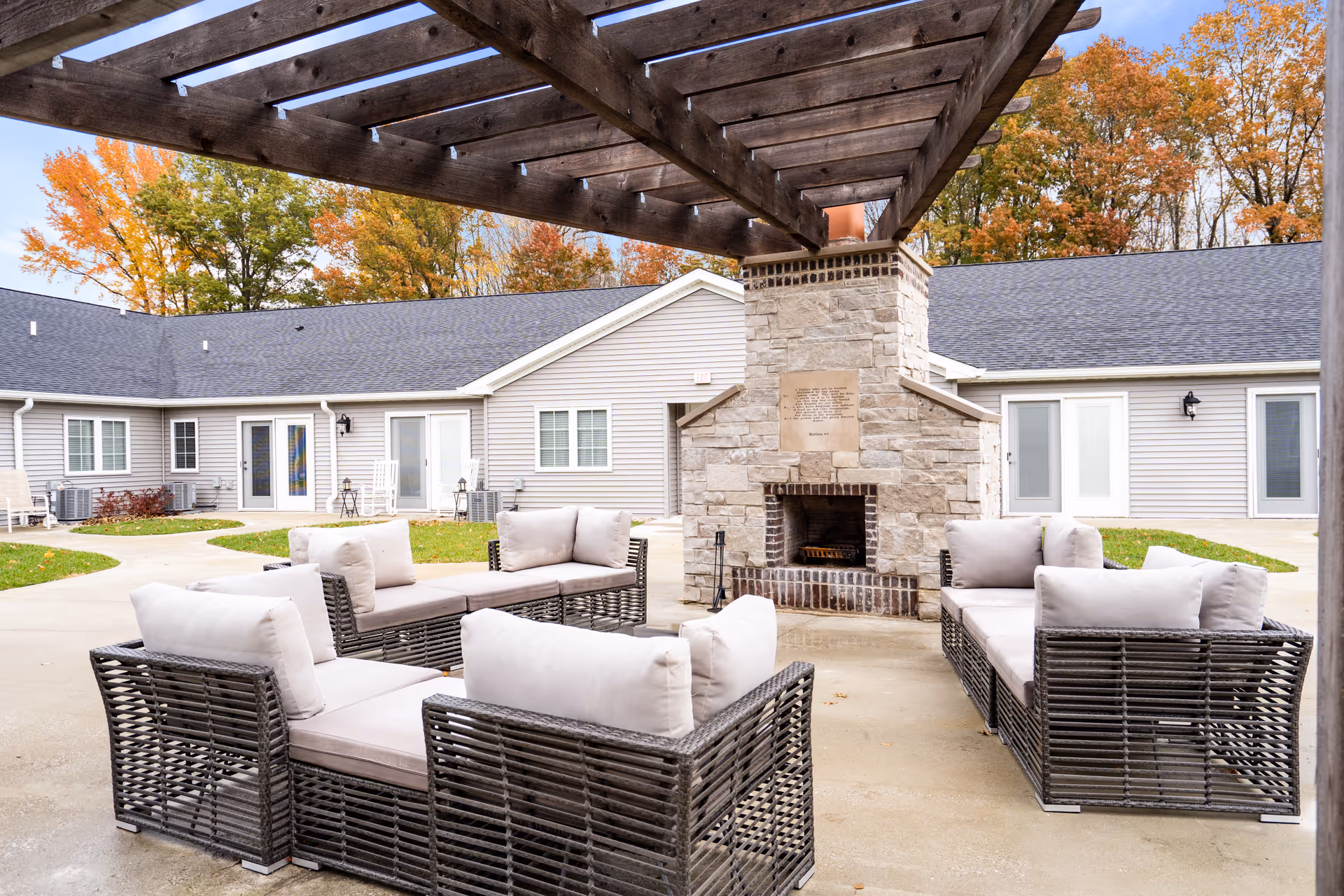 Outdoor seating area with cushioned wicker sofas arranged around a stone fireplace under a wooden pergola, with a single-story building and autumn trees in the background.