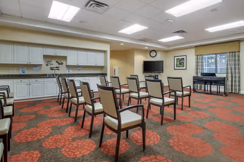 A room with multiple chairs arranged in rows facing a television mounted on the wall. The room has a patterned carpet with red and brown colors, white cabinets along one wall, a clock above the television, a framed picture on the wall, and a piano near a window with curtains.