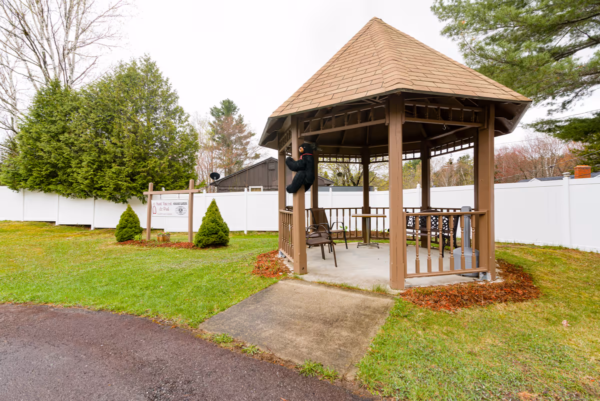 A brown wooden gazebo with a shingled roof situated on a grassy lawn. Inside the gazebo are several chairs and a small table. A black bear decoration is hanging on one of the gazebo's posts. In the background, there is a white fence and a sign that reads 'St. Vincent de Paul Rehabilitation and Nursing Center' surrounded by small shrubs and trees.