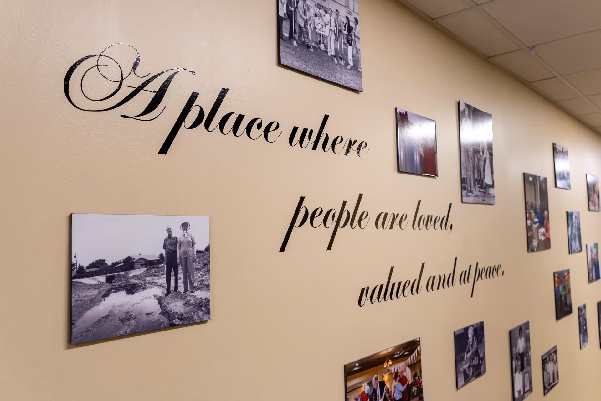 A beige wall decorated with black cursive text that reads 'A place where people are loved, valued and at peace,' surrounded by various framed black and white and color photographs depicting people and activities.