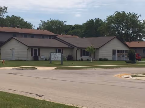 Exterior of a single-story assisted living facility in Monroe, Wisconsin, with a sign and parking.