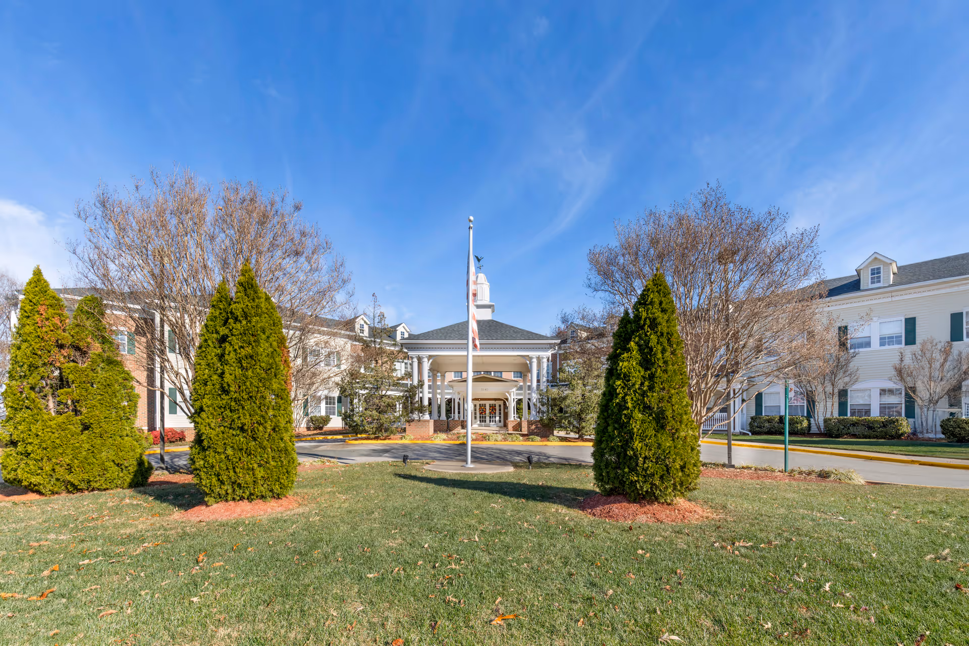 Front exterior view of Brookdale Lake Ridge facility with a circular driveway, flagpole, manicured lawn, and several trees under a clear blue sky.