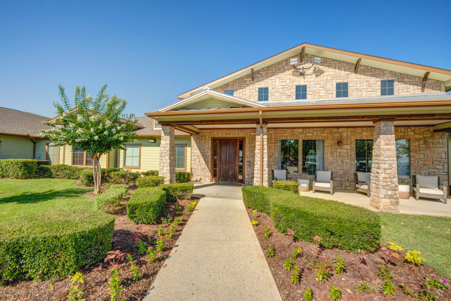 Front exterior view of Santa Fe Trails Assisted Living and Memory Care building with stone facade, a covered entrance supported by stone pillars, neatly trimmed bushes, flower beds, and a clear blue sky.