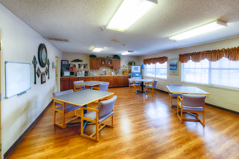 A bright room with wooden flooring and several tables each surrounded by chairs. The room has large windows with valance curtains letting in natural light. Along one wall, there is a kitchenette area with wooden cabinets, a microwave, and decorative items. A whiteboard and wall clock are mounted on another wall.