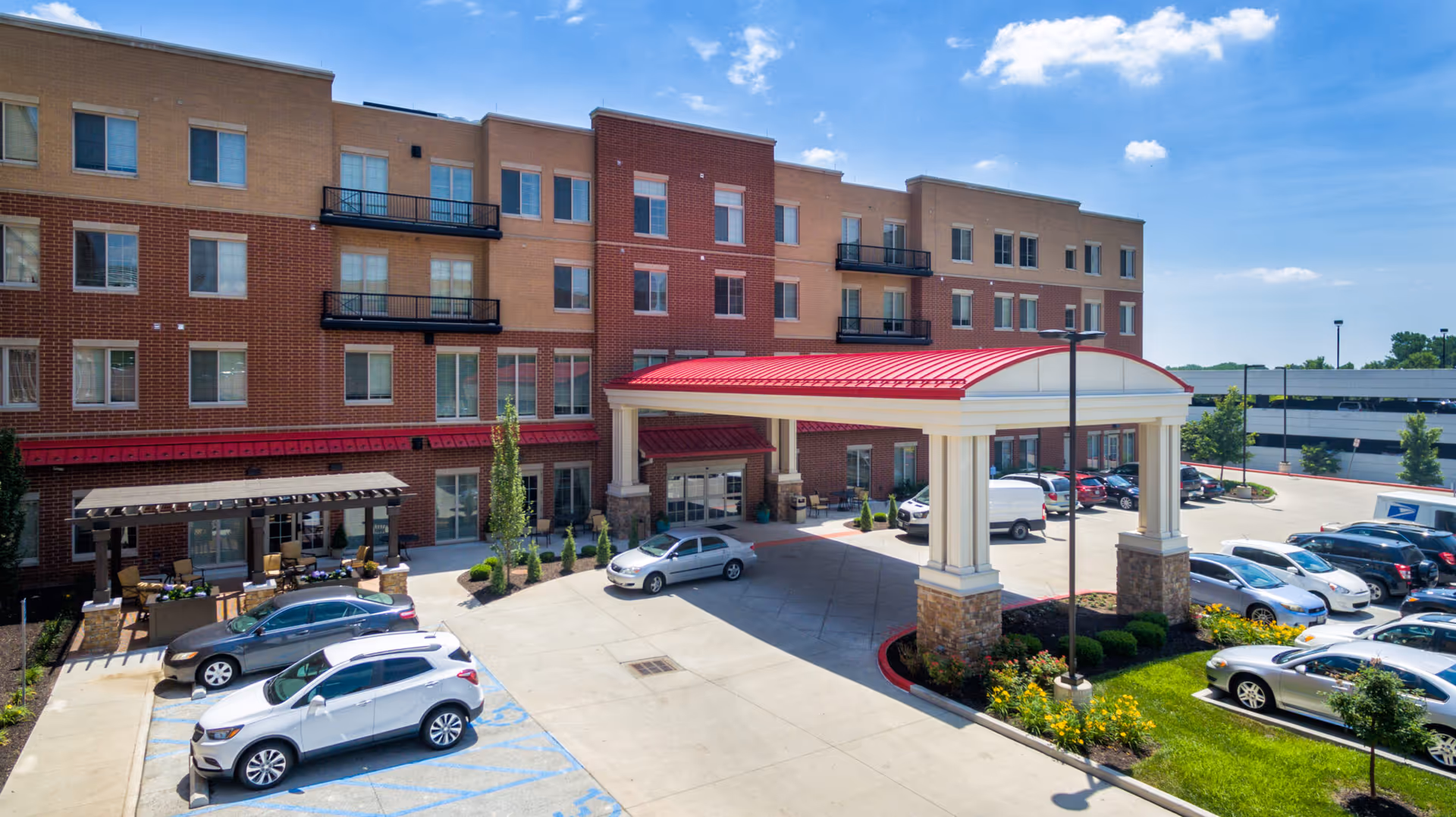 Exterior view of a multi-story assisted living facility with a red roofed entrance canopy, several parked cars, and a small landscaped area with flowers and shrubs under a blue sky with some clouds.