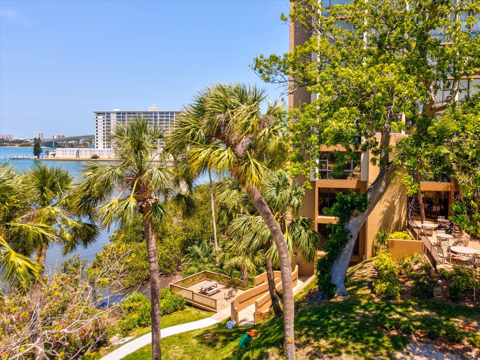 Outdoor view of a senior living facility with palm trees, a wooden picnic table area, a walking path, and a building with large windows and outdoor seating. Water and another building are visible in the background under a clear blue sky.