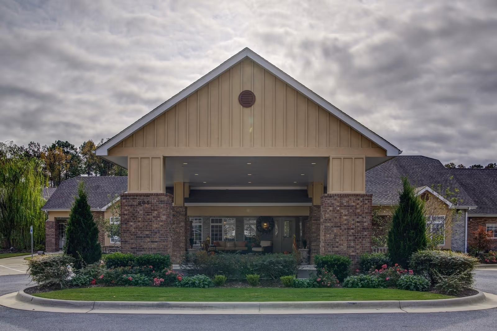 Front exterior view of Brookfield Assisted Living facility showing a covered entrance with brick pillars, landscaped greenery, and a cloudy sky overhead.