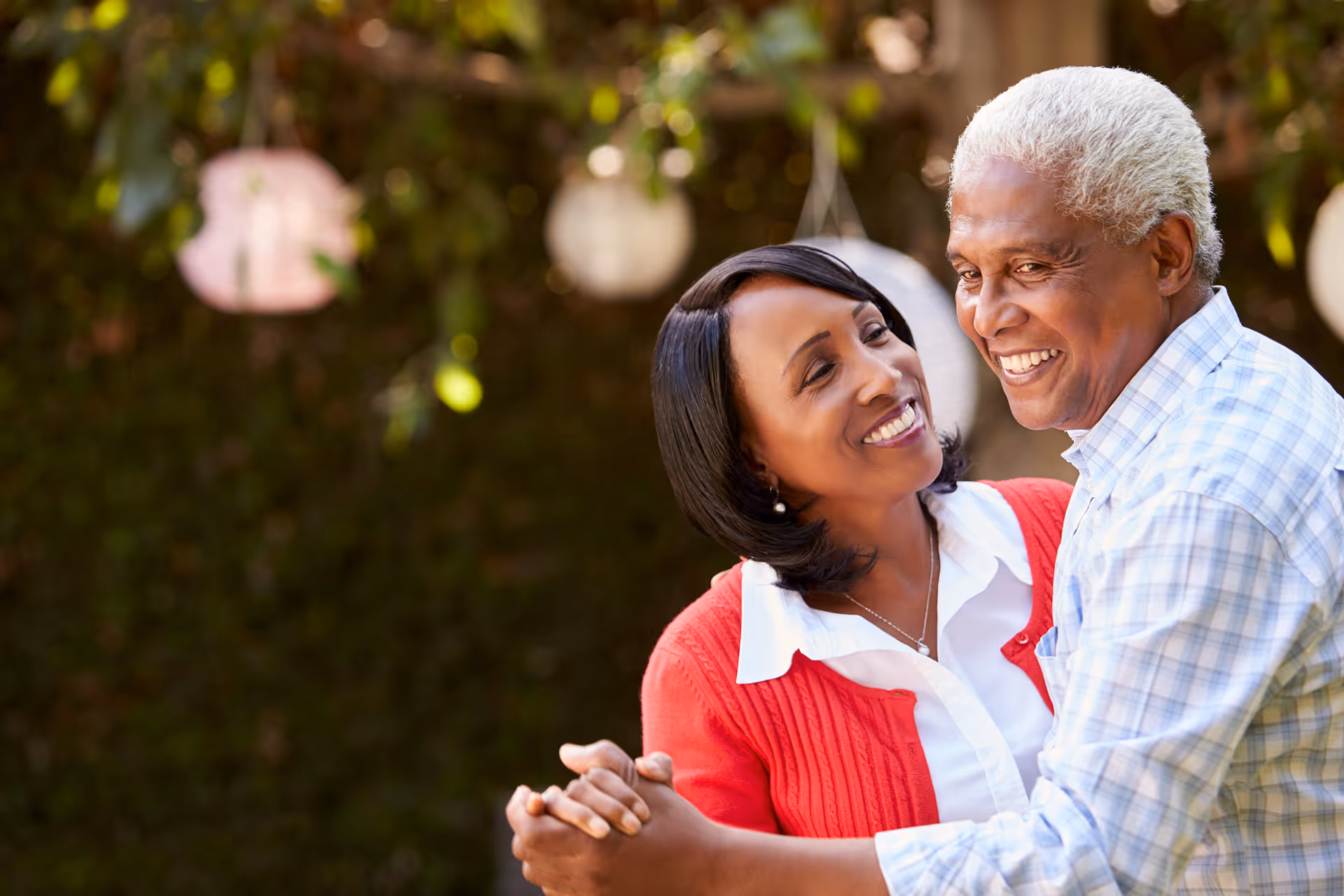 An elderly couple smiling and dancing together outdoors with greenery and hanging lanterns in the background.