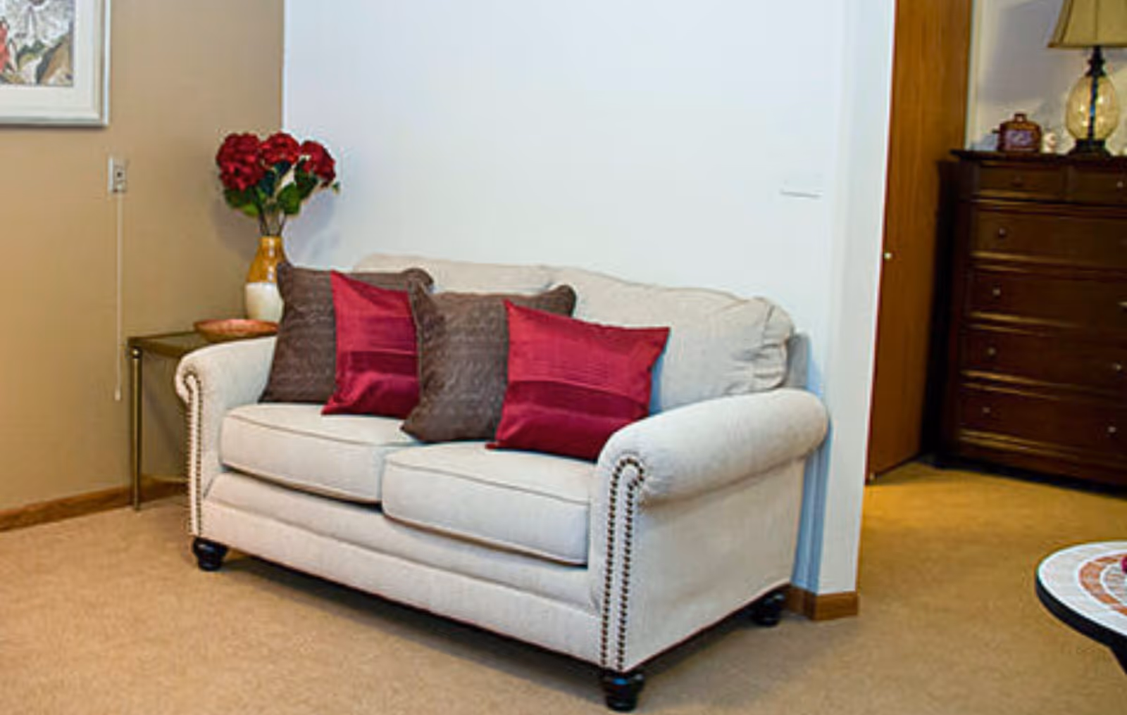 A beige upholstered sofa with four decorative pillows, two in dark brown and two in deep red, placed against a white wall. To the left of the sofa is a small side table with a vase of red flowers. To the right, there is a wooden dresser with a lamp and decorative items on top. The room has beige carpet flooring.