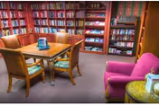 Cozy library-style room with bookshelves lining the walls, a wooden table with four chairs, and a pink upholstered armchair.