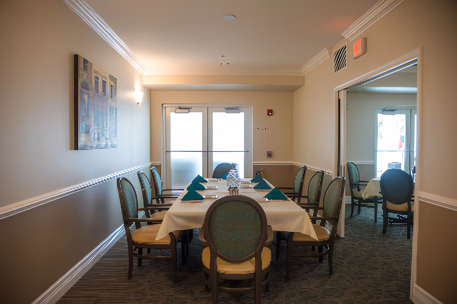 A dining room with a long rectangular table covered with a white tablecloth and set with teal folded napkins and silverware. The room has beige walls with a painting on the left wall and double glass doors at the far end. There are several wooden chairs with teal upholstered backs and beige seats arranged around the table. A mirror on the right wall reflects part of the room and another table with chairs.