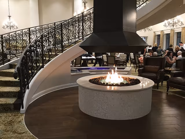 Interior view of a senior living facility lounge area featuring a central round fireplace with a black hood, surrounded by seating including armchairs. In the background, there is a curved staircase with ornate wrought iron railings and several people seated at tables, engaged in conversation. Elegant chandeliers hang from the ceiling, adding to the warm and inviting atmosphere.