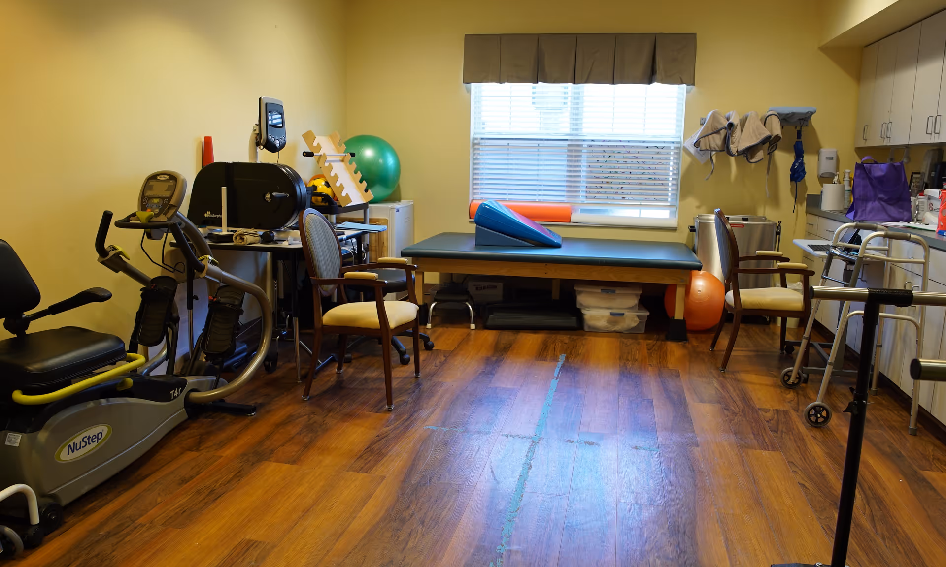 A therapy or rehabilitation room with exercise equipment including a NuStep recumbent cross trainer, therapy table, chairs, exercise balls, walker, and various therapy tools. The room has wooden flooring, yellow walls, a window with blinds, and cabinets along one wall.