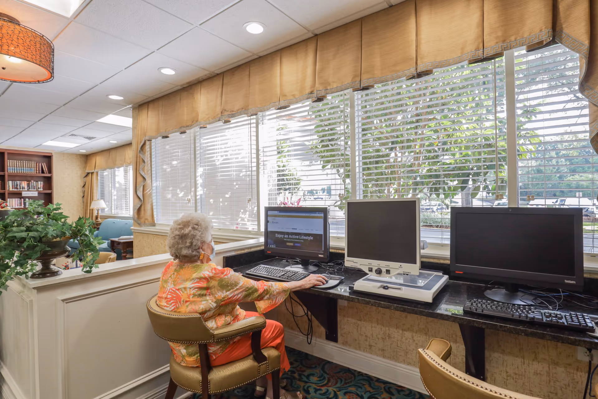 An elderly woman wearing a floral shirt and orange pants sits at a computer workstation in a well-lit room with large windows covered by blinds. The workstation has three computer monitors and keyboards. The room has beige walls, a patterned carpet, and a bookshelf with books and a lamp in the background.