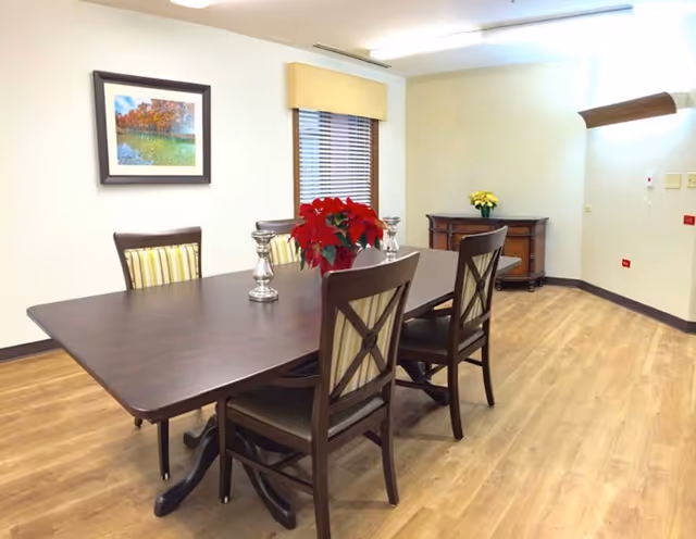 A dining room with a long dark wood table, four chairs, a red poinsettia centerpiece, wood flooring, and framed art on the wall.