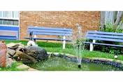A small landscaped pond with a central fountain, rocks and grass, with blue benches and a brick wall in the background.