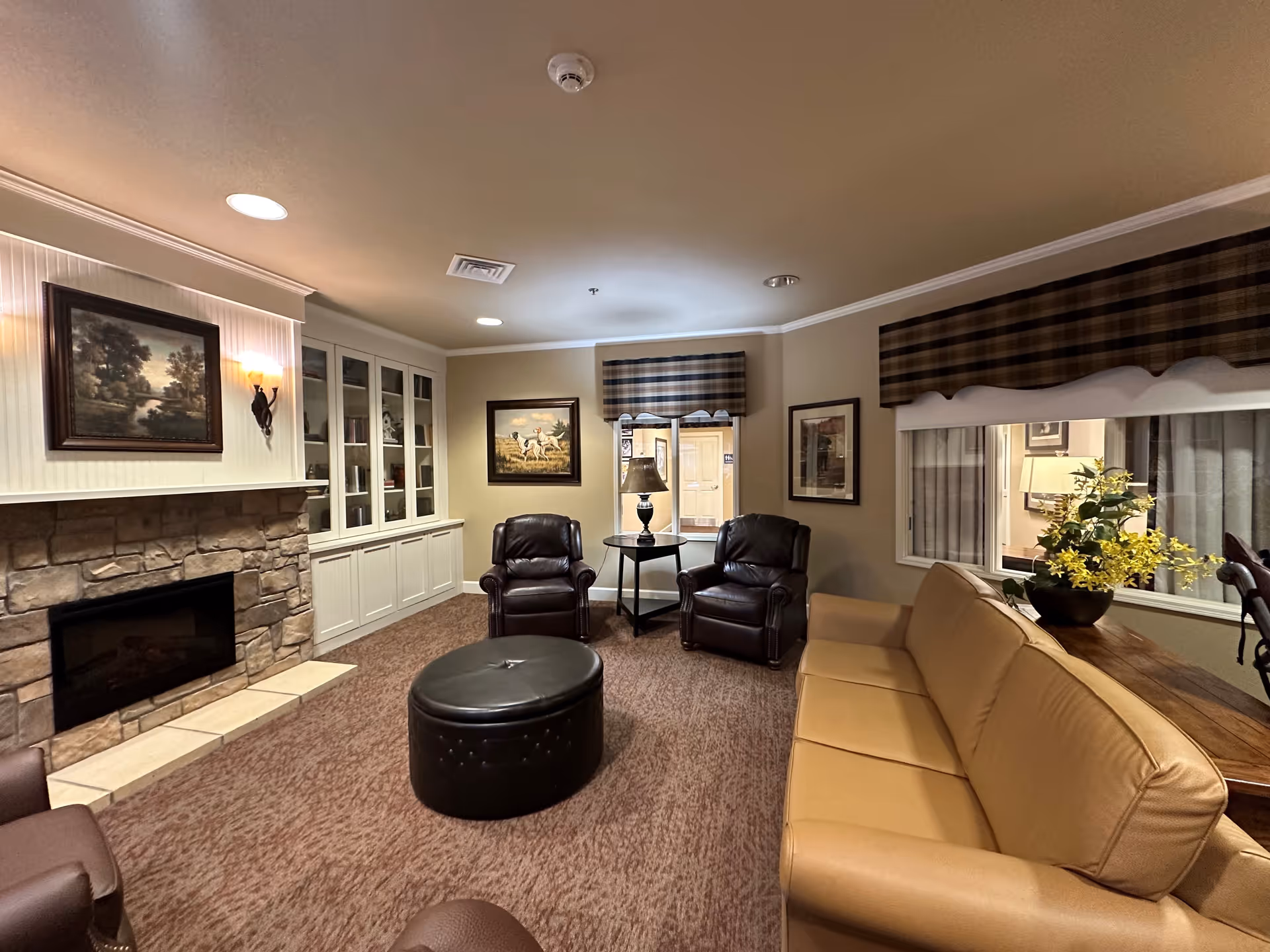 A cozy living room with a stone fireplace on the left wall, a tan leather sofa on the right, two dark leather armchairs facing the fireplace, a round black ottoman in the center, and a side table with a lamp between the armchairs. The walls are decorated with framed paintings and windows with plaid valances. There is a built-in white cabinet with glass doors on the left side of the room.