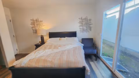 A bedroom with a large bed featuring a striped comforter and pillows. There is a nightstand with a lamp on the left side of the bed and a dark upholstered chair on the right. Two decorative metal wall art pieces hang above the bed. A sliding glass door on the right side opens to an outdoor area.