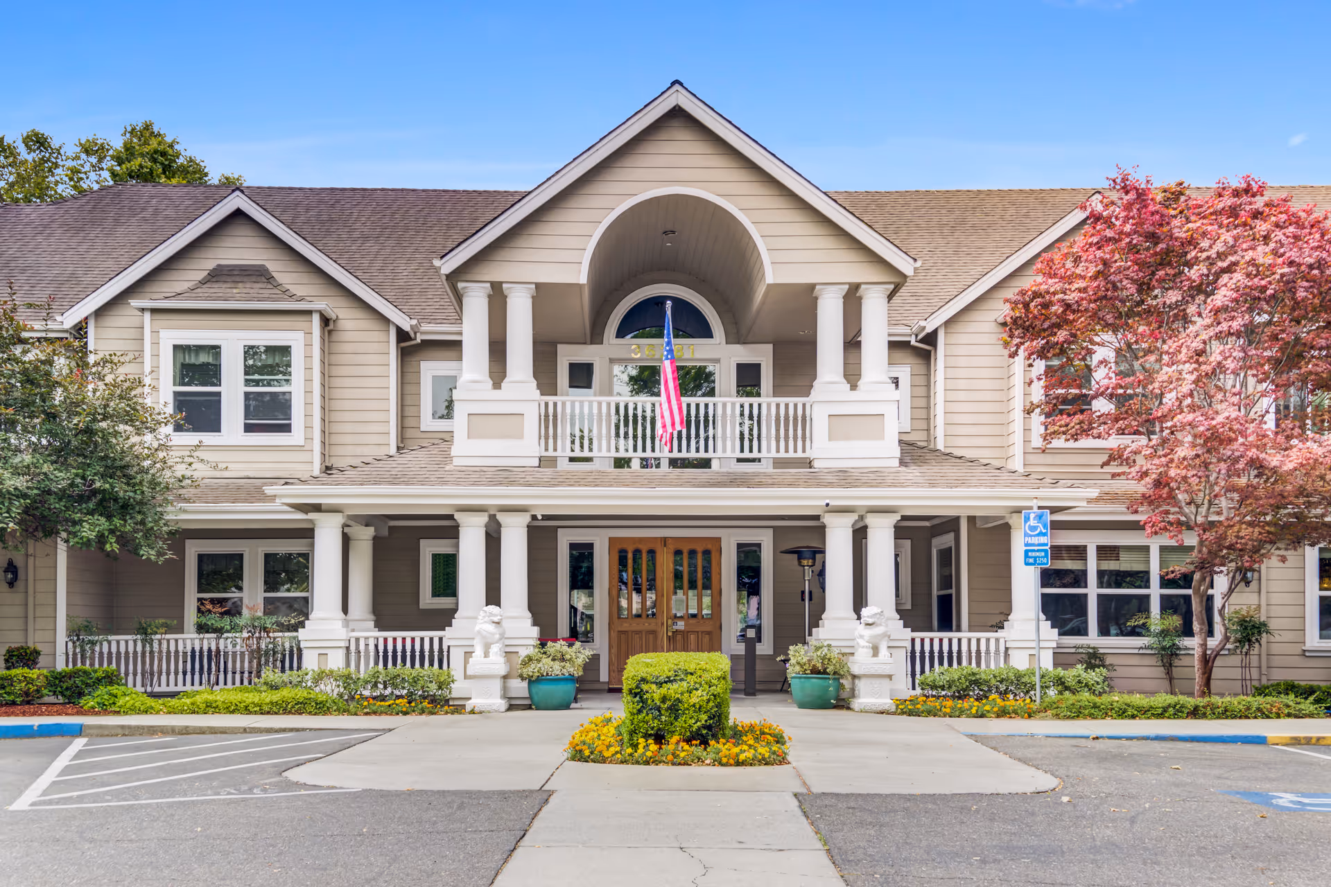 Front exterior view of a two-story senior living facility building with beige siding, white columns, and a covered entrance. There is an American flag hanging above the entrance, two white lion statues on either side of the walkway, and landscaped greenery including bushes and trees with red leaves. Handicap parking spaces are visible in front of the building.