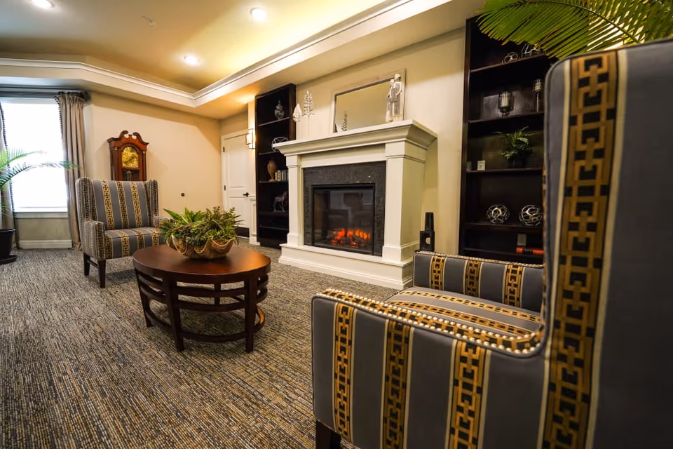 A cozy living room area with two patterned armchairs, a wooden coffee table with a plant centerpiece, a fireplace with a white mantel, built-in dark wood shelves with decorative items, a grandfather clock, and a window with curtains allowing natural light.