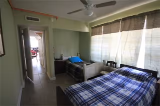 A bedroom with two single beds, one with a blue plaid blanket and the other with a dark plaid blanket. There is a ceiling fan above, a window with closed blinds on the right, and a doorway leading to another room on the left.