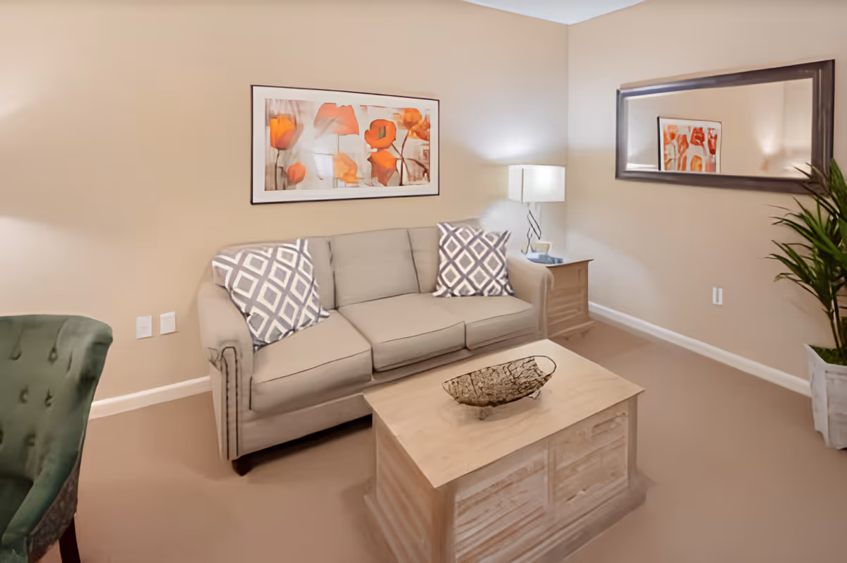 A cozy living room with a beige sofa adorned with two patterned pillows, a wooden coffee table with a decorative bowl, a side table with a lamp, a large wall mirror, a framed floral artwork, and a potted plant in the corner.