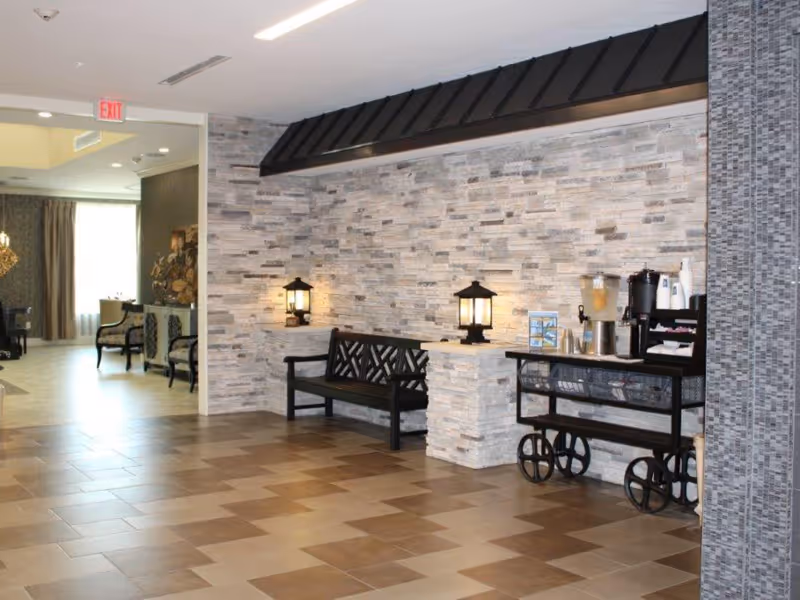 Interior view of a senior living facility with a tiled floor and a stone accent wall. There is a black bench with decorative backrest and two lantern-style lamps on a stone ledge. To the right, a black metal cart holds a beverage dispenser, coffee maker, cups, and other supplies. In the background, there are chairs and a window with curtains allowing natural light to enter.