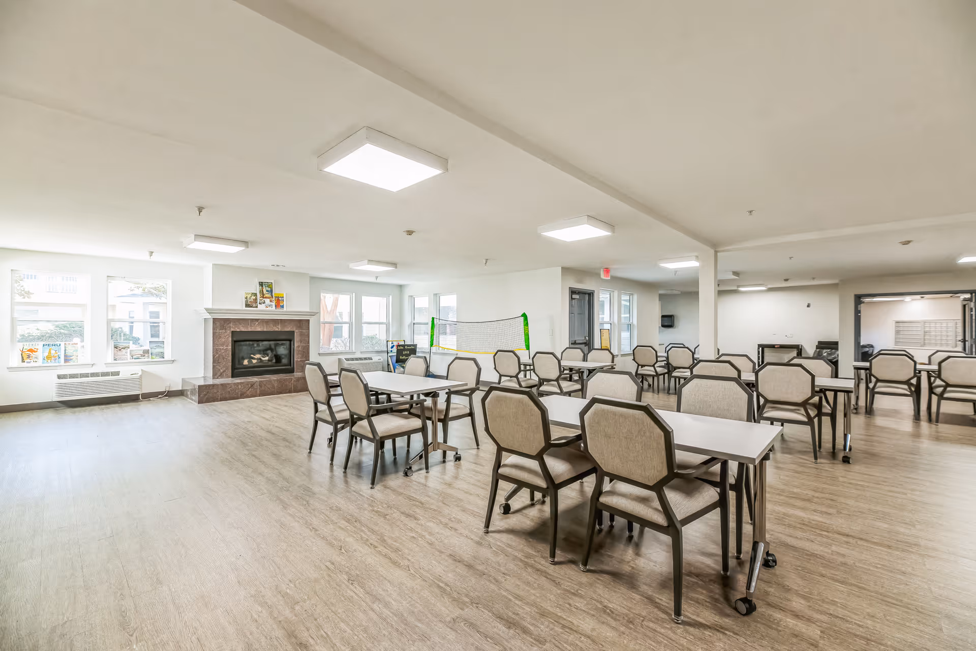 Large bright common dining/activity room with multiple tables and chairs, a fireplace, and windows.