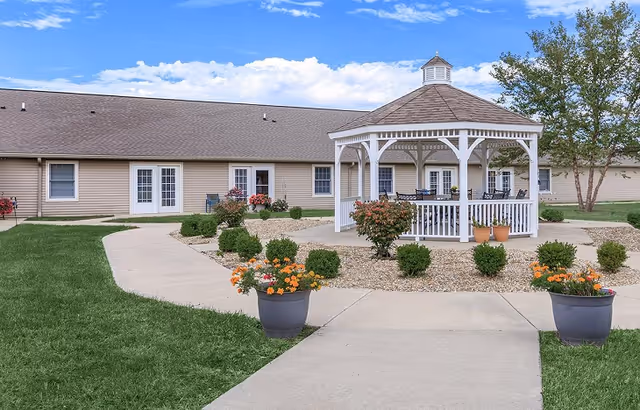 Outdoor area of Rochester Estates Senior Living featuring a white gazebo surrounded by small bushes and flower pots with orange and yellow flowers. The gazebo is situated on a gravel bed with a concrete walkway leading to it. In the background, there is a beige building with multiple doors and windows under a partly cloudy blue sky.