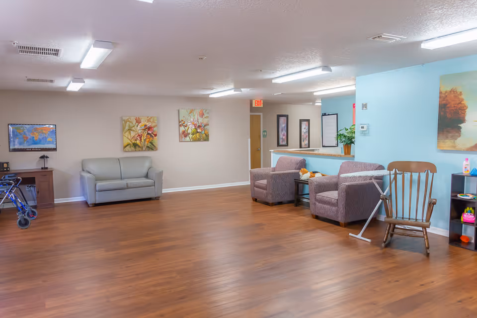 A spacious senior living facility common area with wood flooring, a gray loveseat, two purple armchairs, a wooden rocking chair, and a small black shelf with colorful items. The walls are decorated with floral and landscape paintings. There is a walker near a wooden desk with a lamp and a world map on the wall. The ceiling has multiple fluorescent lights and an exit door is visible in the background.