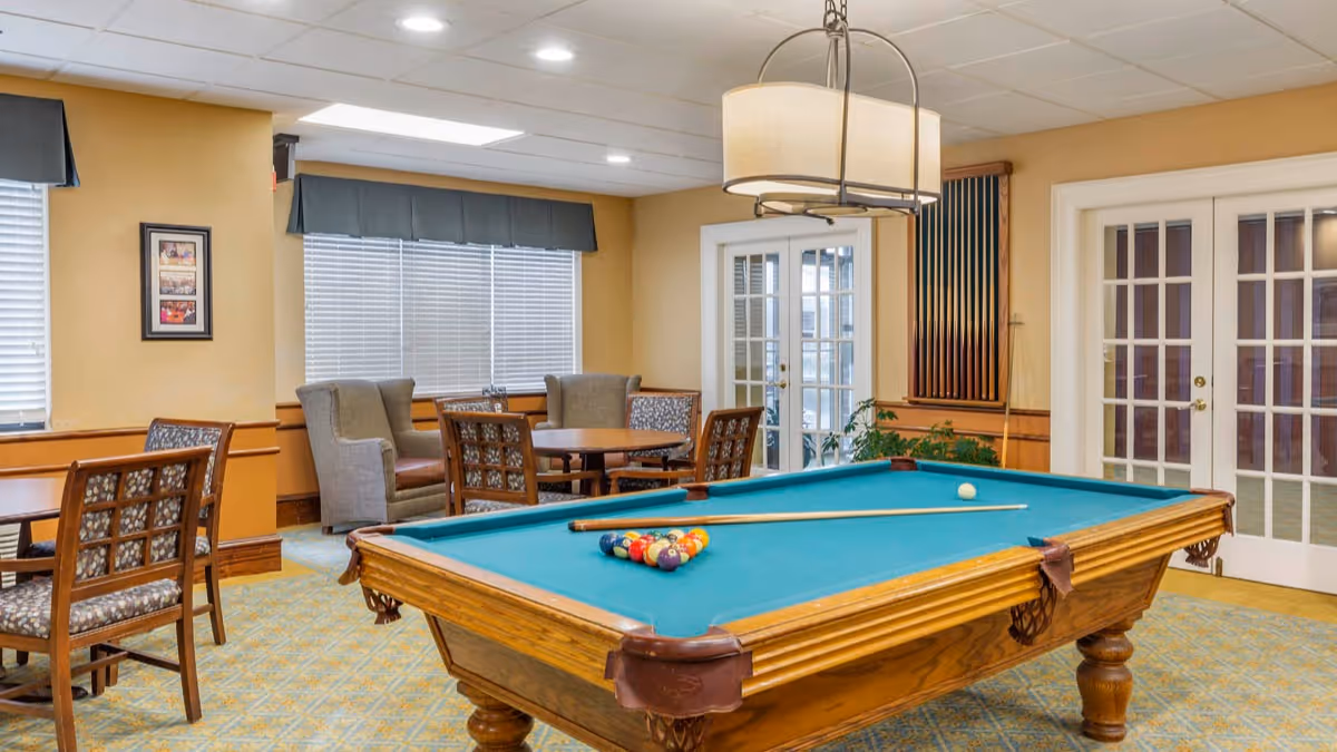 Pool table with racked balls and a cue in a well-lit common room with tables, chairs, and large windows.