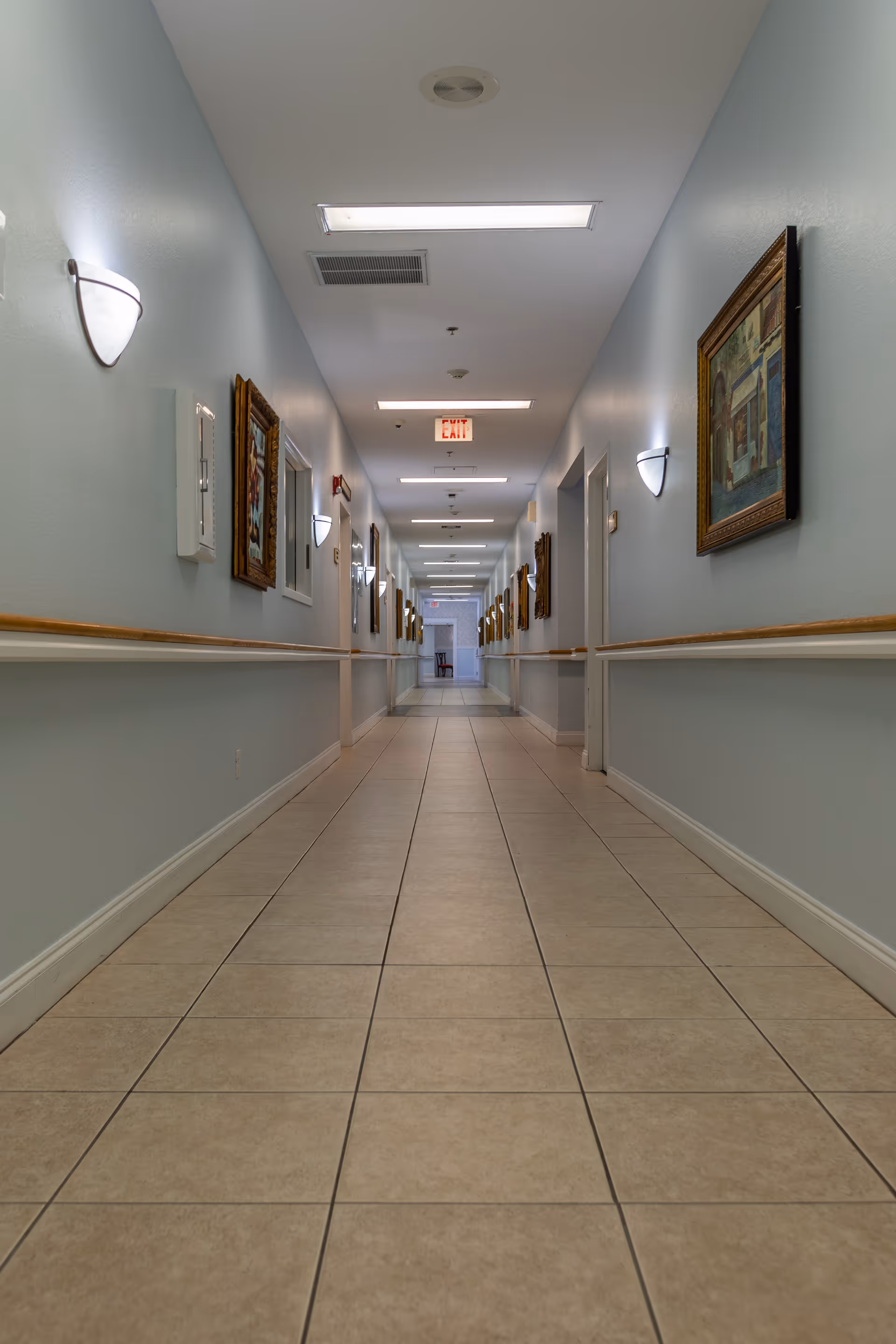 A long, clean hallway with tiled floors and light blue walls, decorated with framed paintings on both sides. The hallway is well-lit with ceiling lights and wall sconces, and there are handrails along the walls. Doors line the hallway, and an exit sign is visible at the far end.